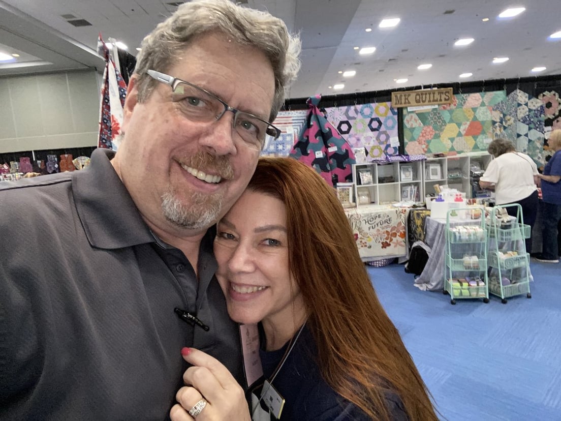 Melissa and Paul (MK Quilts) together at a quilt show, with their booth and quilts on display behind them